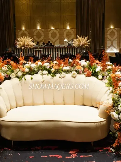 A beautifully designed seating area for honored guests or as a photo spot. The plush white sofa is framed by a cascade of orange and white flowers, creating a vibrant and luxurious focal point.
