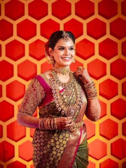 A happy bride against a vibrant backdrop. Her olive green and pink Kanchipuram saree is draped to highlight the rich border and intricate work, ensuring she looks flawless from every angle.