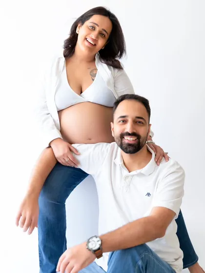A fun and happy couple's maternity portrait with the mother-to-be seated above her partner. This pose creates a playful and connected image in a bright studio setting.