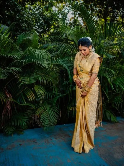 A full-length shot of the bride, her golden saree draped elegantly against a backdrop of lush palm trees.
