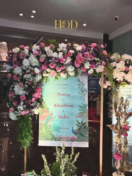 A close-up of a personalized wedding welcome sign featuring beautiful peacock illustrations, a nod to traditional Indian artistry, framed by a lush floral arch.