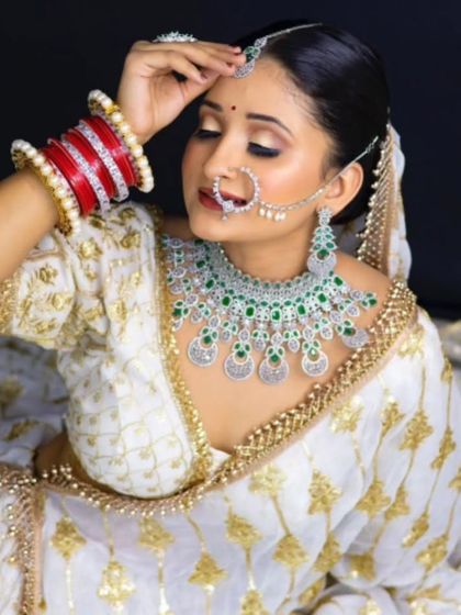 A bride in a white and gold lehenga wearing a beautiful AD and emerald necklace. The contrast of green against the white outfit is striking.