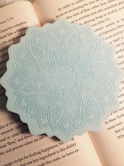 Our Mandala coaster in a calming icy blue color, placed inside a book. The intricate design feels both modern and timeless.