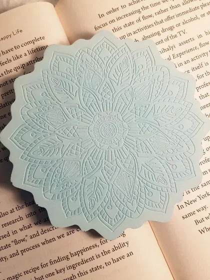Our Mandala coaster in a calming icy blue color, placed inside a book. The intricate design feels both modern and timeless.