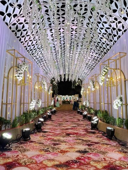 This beautiful entrance passageway is lined with gold-framed arches and features a ceiling with intricate latticework and hanging white flowers. The patterned carpet and spotlights complete this stunning entryway.