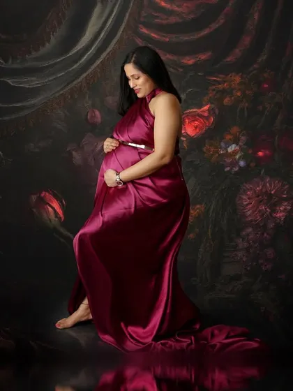 A quiet, introspective pose in a deep magenta satin gown. The painterly floral background and reflective floor add layers of depth and artistry to this beautiful maternity portrait.