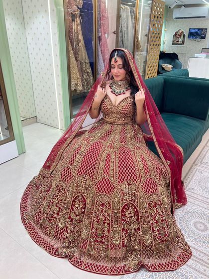 A seated portrait showcasing the magnificent flare and detailed craftsmanship of this red bridal lehenga. The golden zardozi work truly stands out against the rich red velvet.