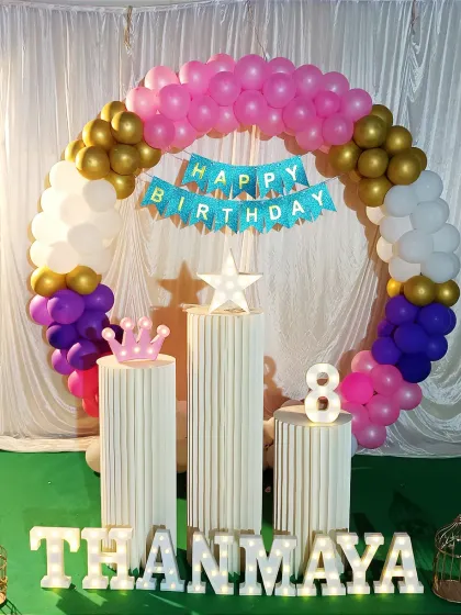 A colorful birthday setup with a circular balloon arch in pink, purple, and gold. The decoration includes a 'Happy Birthday' banner, pedestals, and light-up marquee letters.
