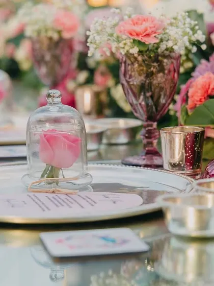A stunning tablescape for a sit-down dinner, featuring silver platters, pink glassware, and a single pink rose preserved under a glass bell jar at each place setting.