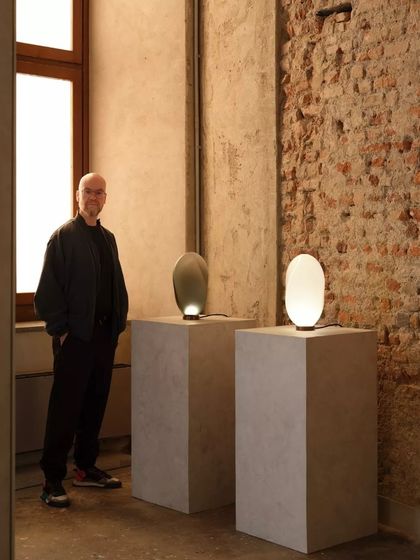 The designer stands beside two Solace table lamps, showcasing the distinct identities of the white and bronze glass versions. This highlights the innovative glassblowing techniques used to create each unique piece.
