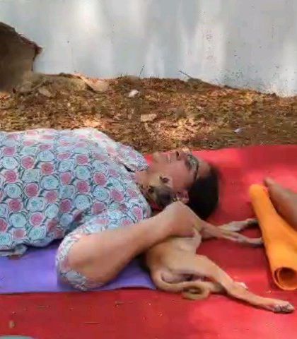 A participant lies in shavasana on a red mat outdoors, with a tiny puppy snuggled up against her neck, completely relaxed.