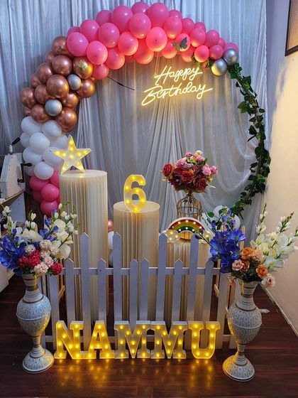 An elegant birthday decoration in pink and rose gold tones. This setup features a semi-circular balloon arch with ivy accents, a neon sign, and a light-up number '6' for the milestone celebration.