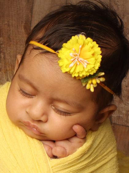 A close-up portrait of a sleeping baby in a vibrant yellow wrap with a matching floral headband. The hand gently resting on the cheek is a classic sign of a comfortable, dreaming baby.