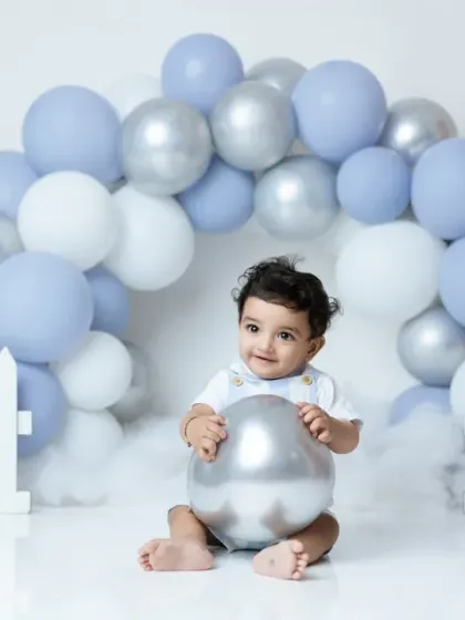 A sweet portrait from a first birthday shoot, with the baby boy sitting under a beautiful arch of blue, white, and silver balloons.