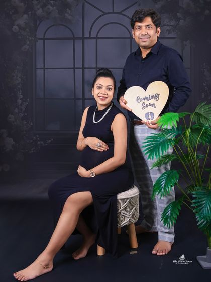 The couple announces their joy with a "Coming Soon" heart prop. This full-length shot showcases their coordinated dark outfits against a moody, elegant backdrop.