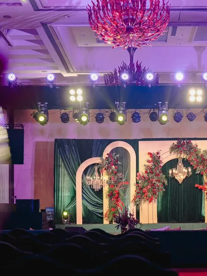The stage for this modern Sangeet featured a backdrop of emerald green velvet and elegant gilded arches, asymmetrically decorated with crimson floral arrangements for a contemporary, artistic feel.