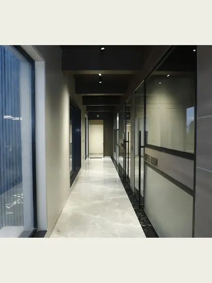 A long corridor in a corporate office, lined with glass-walled rooms. The polished marble flooring and recessed ceiling lights create a clean, bright, and professional pathway, enhancing navigation through the space.