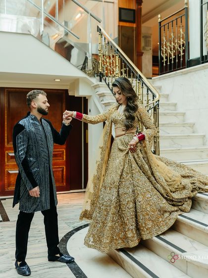 A grand and elegant shot of the couple on a marble staircase, the groom leading his bride down, her golden lehenga flowing beautifully.