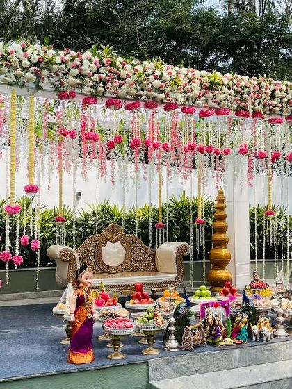 A grand outdoor stage setup perfect for a large-scale Seemantham or engagement. The floral mandapam is decorated with hanging tuberose and pink flower balls, with a beautiful display of traditional offerings in the front.