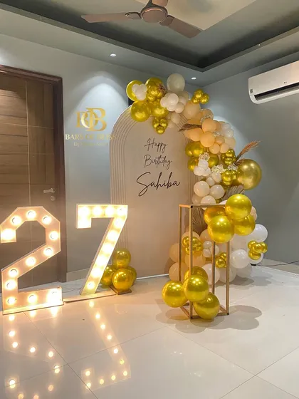 Chic, modern, and personal. This 27th birthday setup features a clean aesthetic with a personalized arched backdrop, a cascade of gold and white balloons, and bold, light-up marquee numbers that create the perfect photo opportunity.