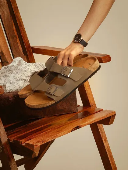 A styled shot of sandals placed on a wooden chair. The warm, earthy tones and the hand reaching for the product create a sense of tactile connection and natural comfort.