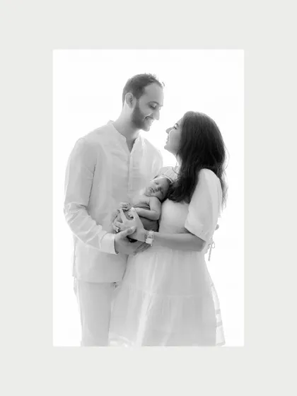 The first family portrait. This black and white image shows new parents gazing at each other with love while securely holding their newborn. The clean, high-key background puts all the emphasis on this new bond and the beginning of their journey as a family of three.