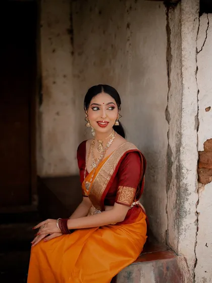 A quiet, seated portrait that radiates traditional elegance. The rustic texture of the wall contrasts beautifully with the smooth silk of the saree and the model's polished look.