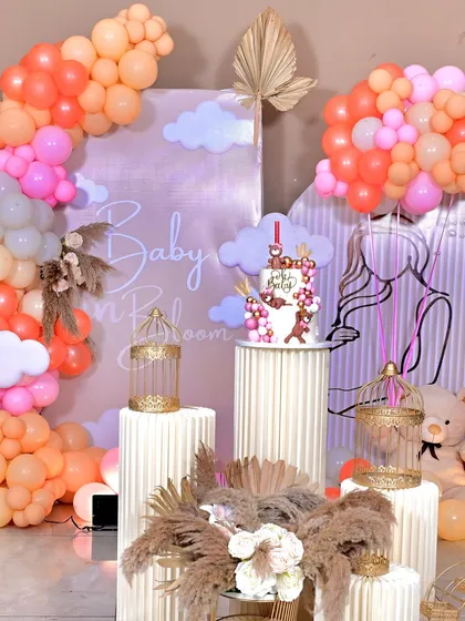 A closer look at our popular 'Baby in Bloom' theme. This setup features a custom backdrop, pampas grass arrangements, themed cake on a fluted pedestal, and a soft, warm color palette of peach and pink balloons.