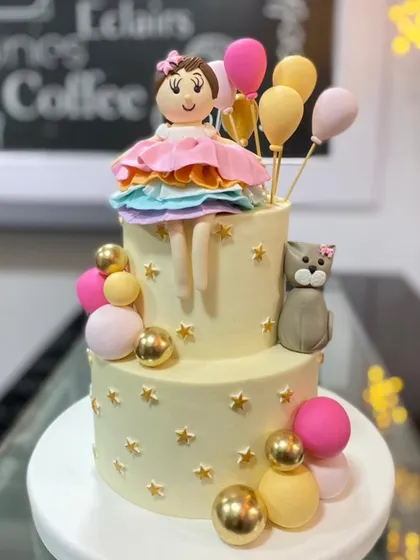 A two-tiered cake with a sweet doll and balloon theme. The design features a handcrafted doll topper, a cute cat, and colorful spheres that look like balloons.