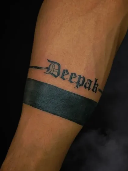 A name tattoo, "Deepak," done in a gothic font over a solid black armband. This creates a bold and personalized statement piece on the forearm.