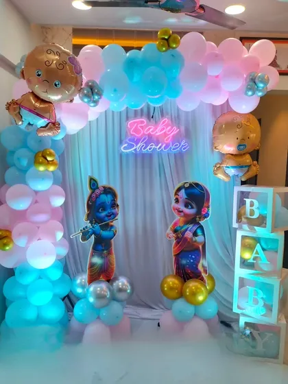 This home baby shower setup combines a pink and blue balloon arch with a 'Baby Shower' neon sign, Radha Krishna cutouts, and transparent 'BABY' blocks.
