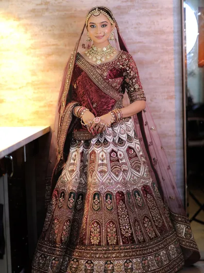 A full view of the bride, standing gracefully. The Polki jewellery set adds a brilliant sparkle that contrasts beautifully with the rich, dark fabric of her outfit.