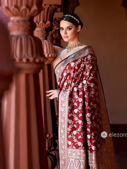 A regal portrait of a model in a deep red, heavily embroidered saree for Elezara. The architectural details and her poised stance evoke the grandeur of Indian palaces.