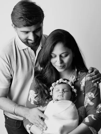 A classic black and white family portrait. The parents look down lovingly at their sleeping newborn, creating a peaceful and emotional image.