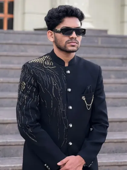 A sharp black Jodhpuri suit with a contemporary twist. It features an abstract peacock feather motif in gold and silver beadwork on the shoulder, paired with a stylish pocket chain.