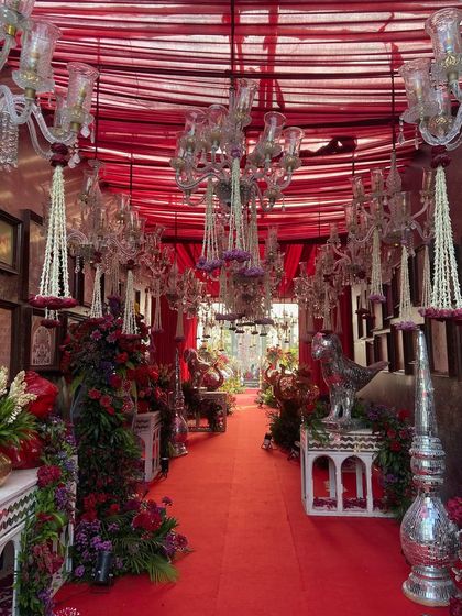 A lavishly decorated passageway for a royal wedding celebration. The design features a ceiling of red drapes, from which hang crystal chandeliers and floral tassels. The red carpet is lined with intricate silver furniture and overflowing red floral arrangements.