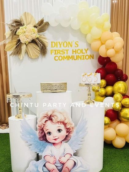 A First Holy Communion decoration for a boy, featuring an angel cutout. The setup uses a warm palette of cream, red, and gold balloons with dried pampas grass florals.