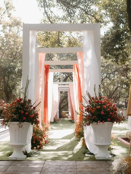 The entrance to the garden ceremony, where guests were welcomed through a modern white archway draped in soft, flowing fabrics and flanked by large urns of vibrant flowers.