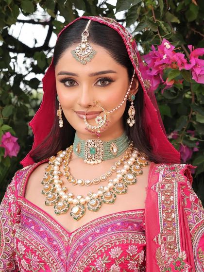 The bride, with her dupatta framing her face, looks divine. The makeup is elevated with a soft glow and rosy tones, perfect against the backdrop of bougainvillea.
