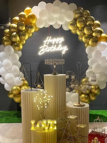 An elegant and classic birthday decoration in gold and white. The full setup includes a circular balloon arch, a neon sign, multiple pedestals with decorative props, and a light-up 'LOVE' sign.