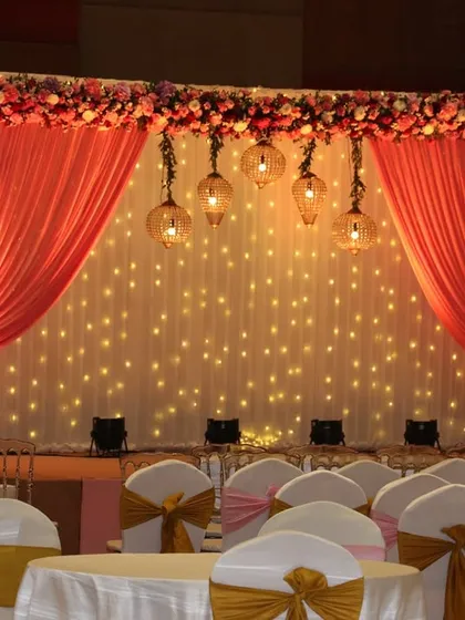 A simple yet elegant stage backdrop with a fairy light curtain, peach-colored drapes, and a thick floral border on top.