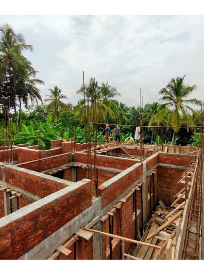 An overview of the ground floor structure of a Kerala residence under construction. You can see the layout of the rooms taking shape with brick walls and concrete beams, surrounded by lush greenery.