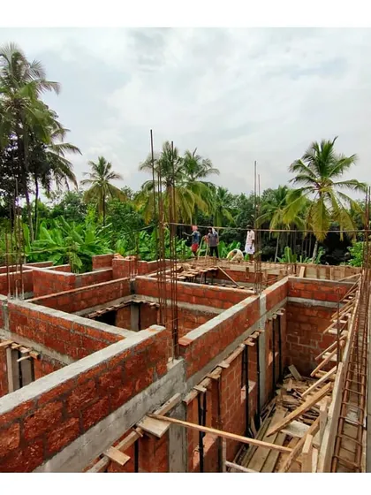 An overview of the ground floor structure of a Kerala residence under construction. You can see the layout of the rooms taking shape with brick walls and concrete beams, surrounded by lush greenery.
