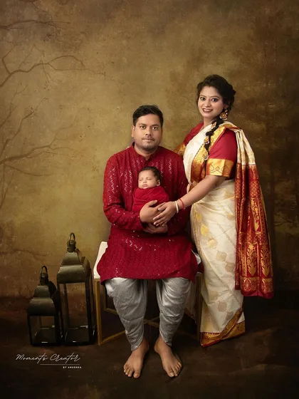 A full-length family portrait in traditional Indian clothing. The father sits holding his newborn while the mother stands beside them, creating a formal yet warm family heirloom.