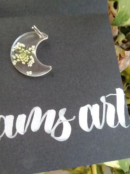 A close-up of the moon-shaped pendant with preserved baby's breath flowers, shown on its custom 'Lams Art' backing card.