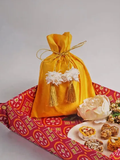 A festive gift set featuring a bright yellow potli bag on a patterned tray. The styling with sweets and flowers makes it look like a perfect, ready-to-give present.