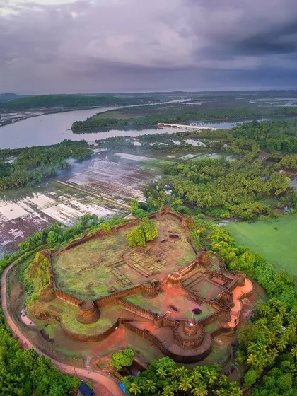 An aerial view of the historic Mirjan Fort near Kumta, enveloped by lush greenery and the winding Aghanashini river. The dramatic monsoon clouds add a layer of mood and history to the scene.