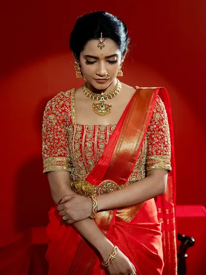 A close-up of a classic South Indian bridal look, featuring a vibrant red Kanjeevaram saree paired with a heavily embroidered blouse. The rich gold zari work and traditional motifs on the blouse showcase the pinnacle of heritage craftsmanship, perfect for a bride seeking authentic grandeur.