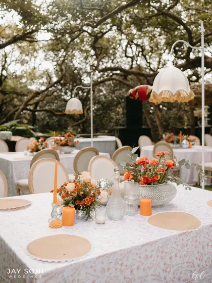 A beautifully set dining table with a floral centerpiece, orange candles, and custom-printed linens, all under the soft glow of a vintage-style lamp.