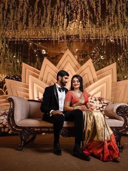 A formally dressed couple is elegantly posed on a luxurious chaise lounge against a grand, lotus-themed stage at their wedding reception. We specialize in capturing the scale and design of large-scale wedding events.